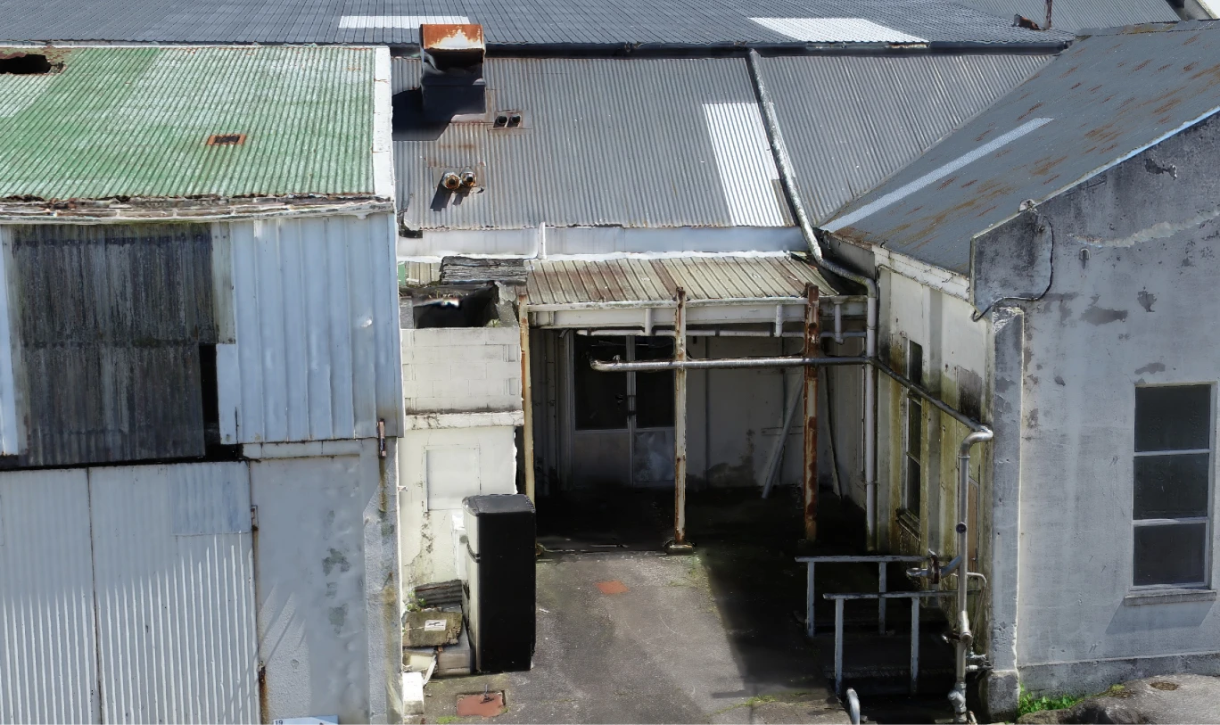 A covered passage between buildings at the Ōkato cheese factory in the 3D inspection model: rusting steel posts holding up a corrugated canopy, pipework running along the wall down to valves, handrails beside steps, a wheelie bin and roof flues behind