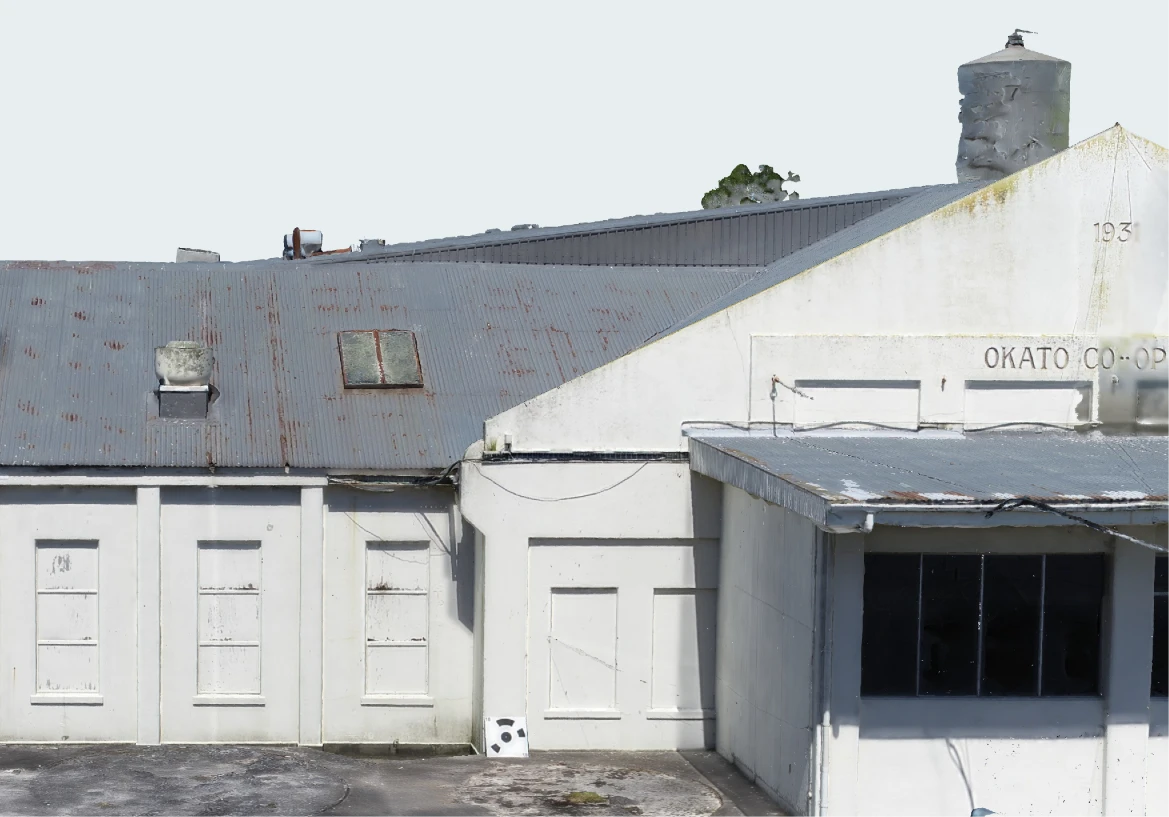 Front of the Ōkato cheese factory in the 3D inspection model: white gabled walls with panelled doors and windows, the lettering OKATO CO-OP on the gable, the water tower behind, and a black-and-white ground control target on the yard