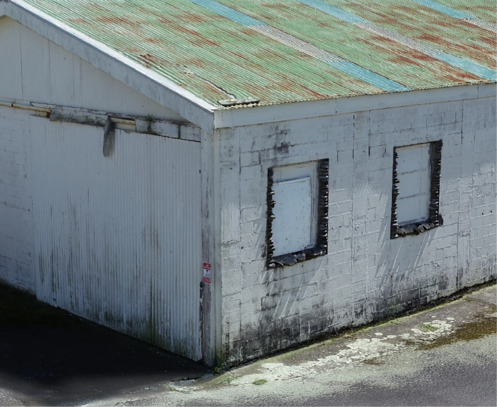 Corner of a building at the Ōkato cheese factory in the 3D inspection model: a rust-streaked corrugated roof, painted block walls with boarded-up windows and weathered frames, a sign by the door, and moss and staining along the foot of the wall