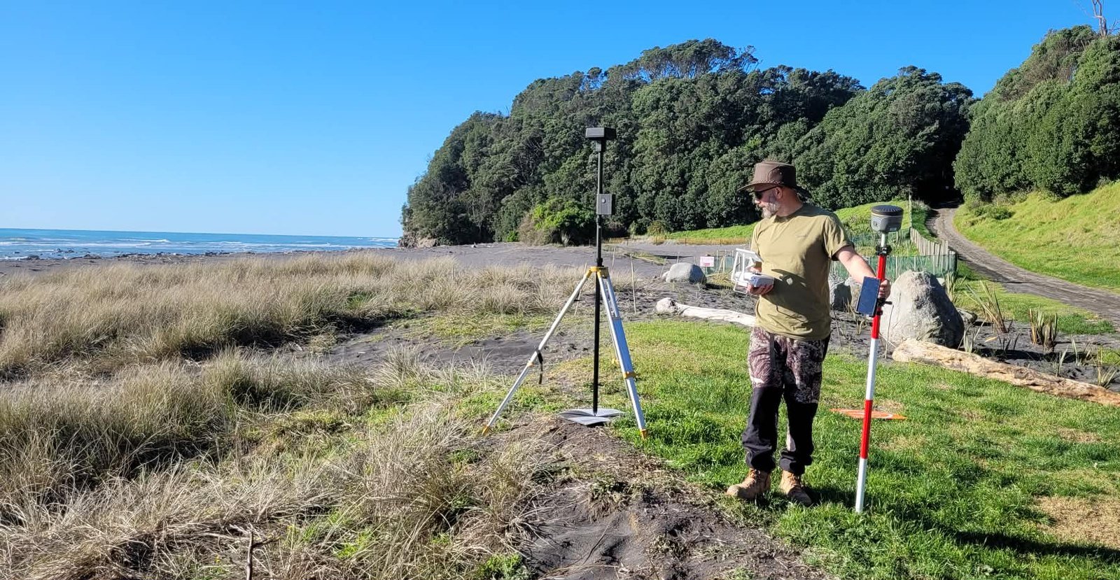 Sam on a grass verge above a black sand beach, holding the drone’s remote controller and the GNSS rover pole, with the DJI D-RTK 2 base station on a tripod beside him