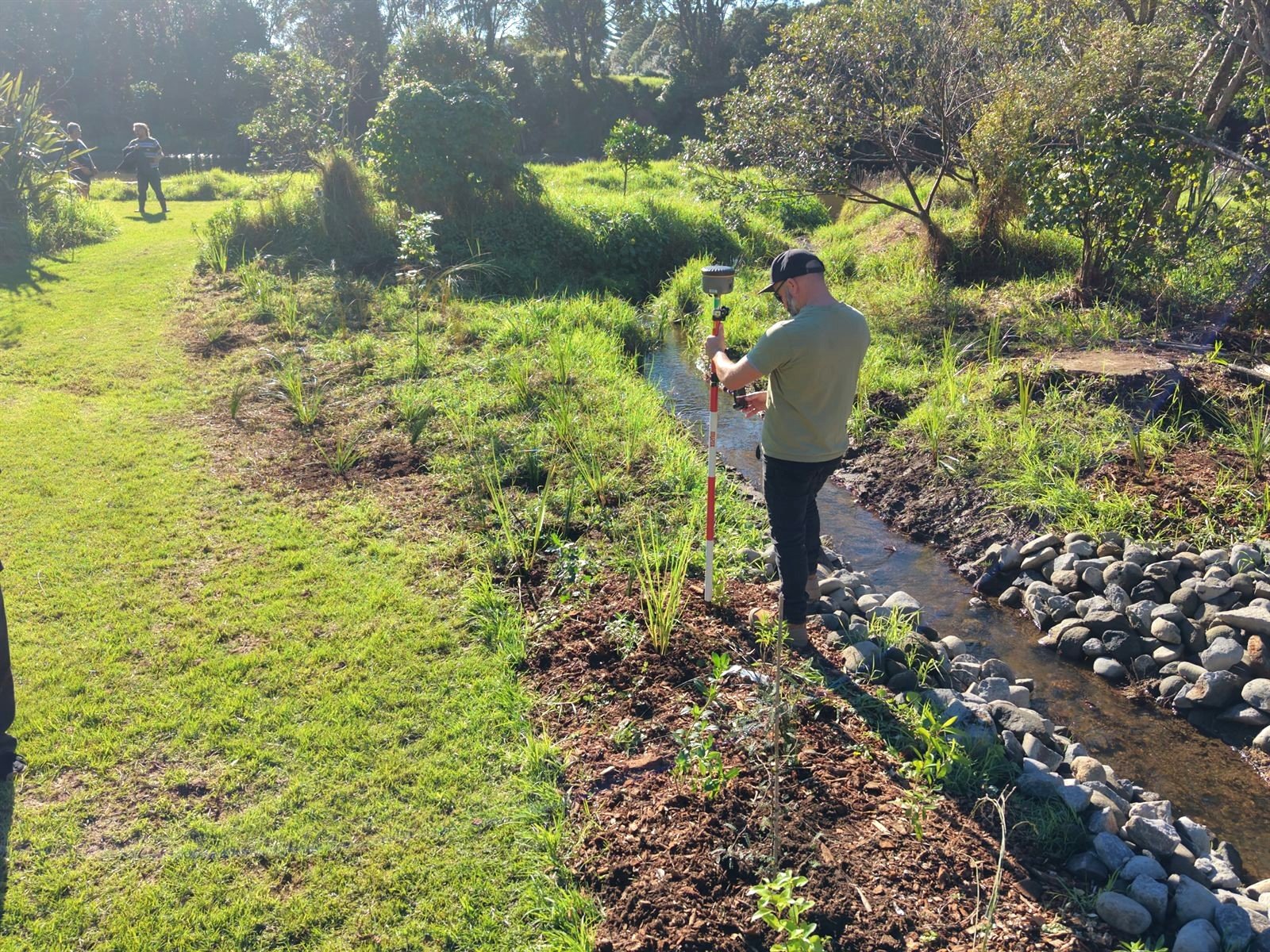 Sam holding the RTK rover pole over a young native plant beside a rock-lined stream in Taranaki