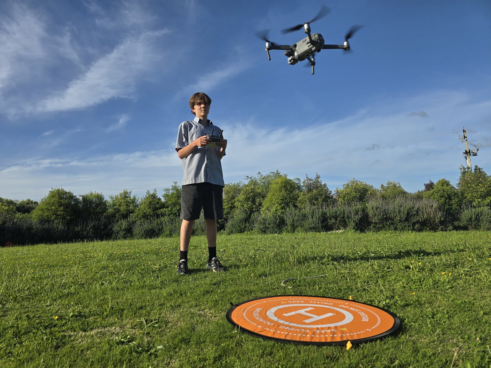 Malachi Kirk holding the controller in a grass paddock, with the drone hovering above an orange launch pad under a blue evening sky