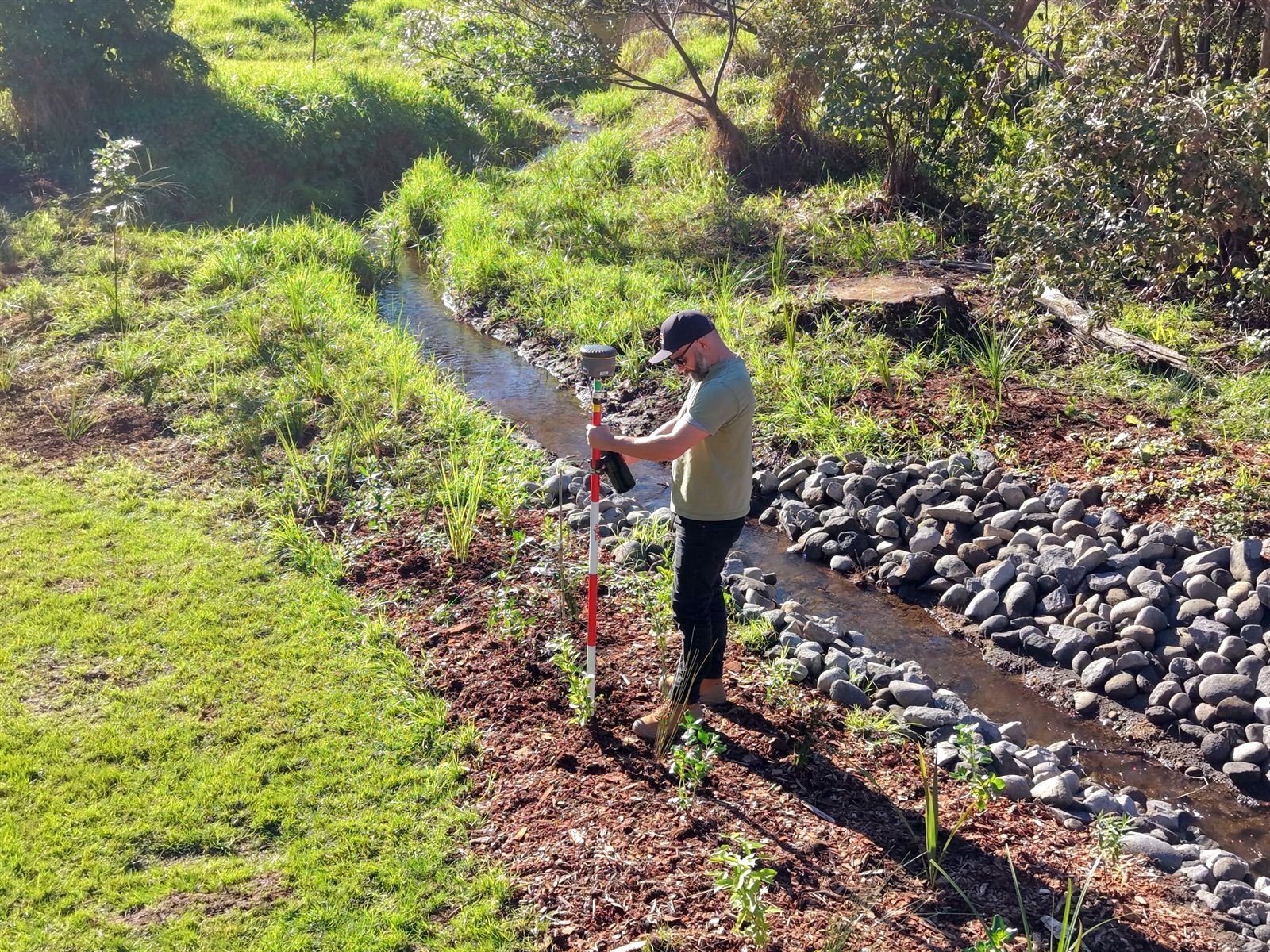 Sam in a cap and sunglasses holding a survey pole with an RTK GNSS receiver over a freshly planted native seedling beside a rock-lined Taranaki stream