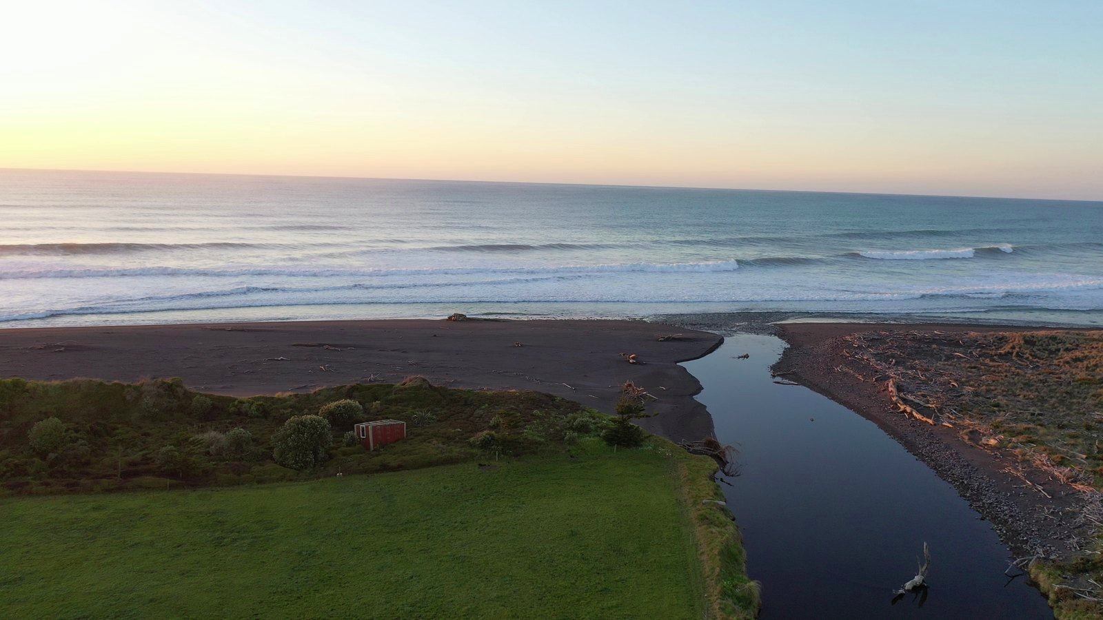 Aerial view at dusk of a Taranaki river mouth meeting the sea, pasture on one side and black sand on the other