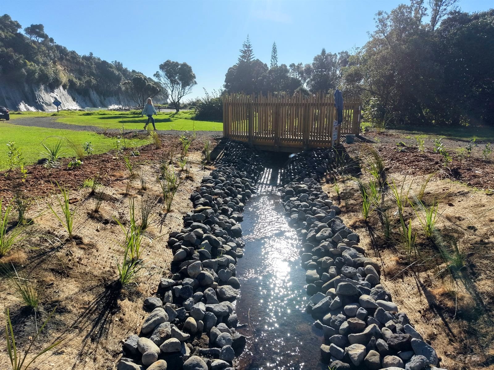 Ground-level view of a newly planted rock-lined stream leading to a timber footbridge, with papa sea cliffs and coast behind on the Taranaki coast